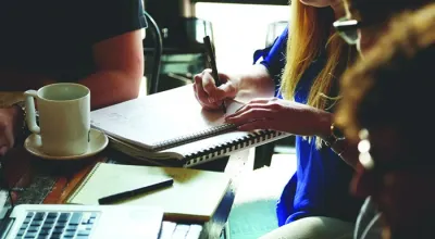 People gathered around a table taking notes in a notebook beside a laptop and coffee cup.
