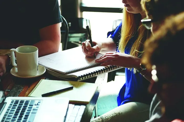 People gathered around a table taking notes in a notebook beside a laptop and coffee cup.